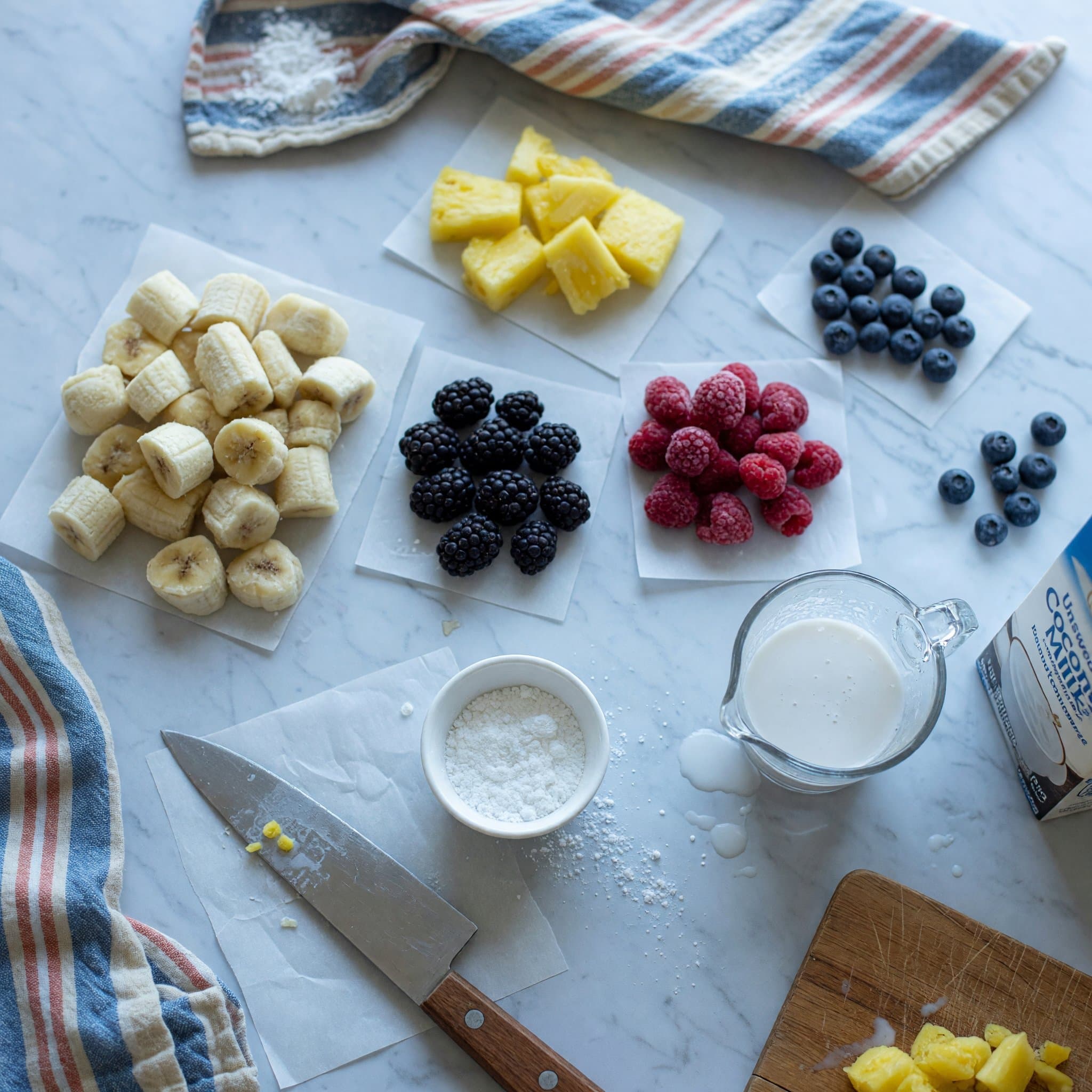 Overhead view of Bananas (), Pineapple, Blackberries, Blueberries, Raspberries, Powdered Sugar and Unsweetened Coconut Milk Beverage ((See Coconut Milk Note Below)) arranged on a table