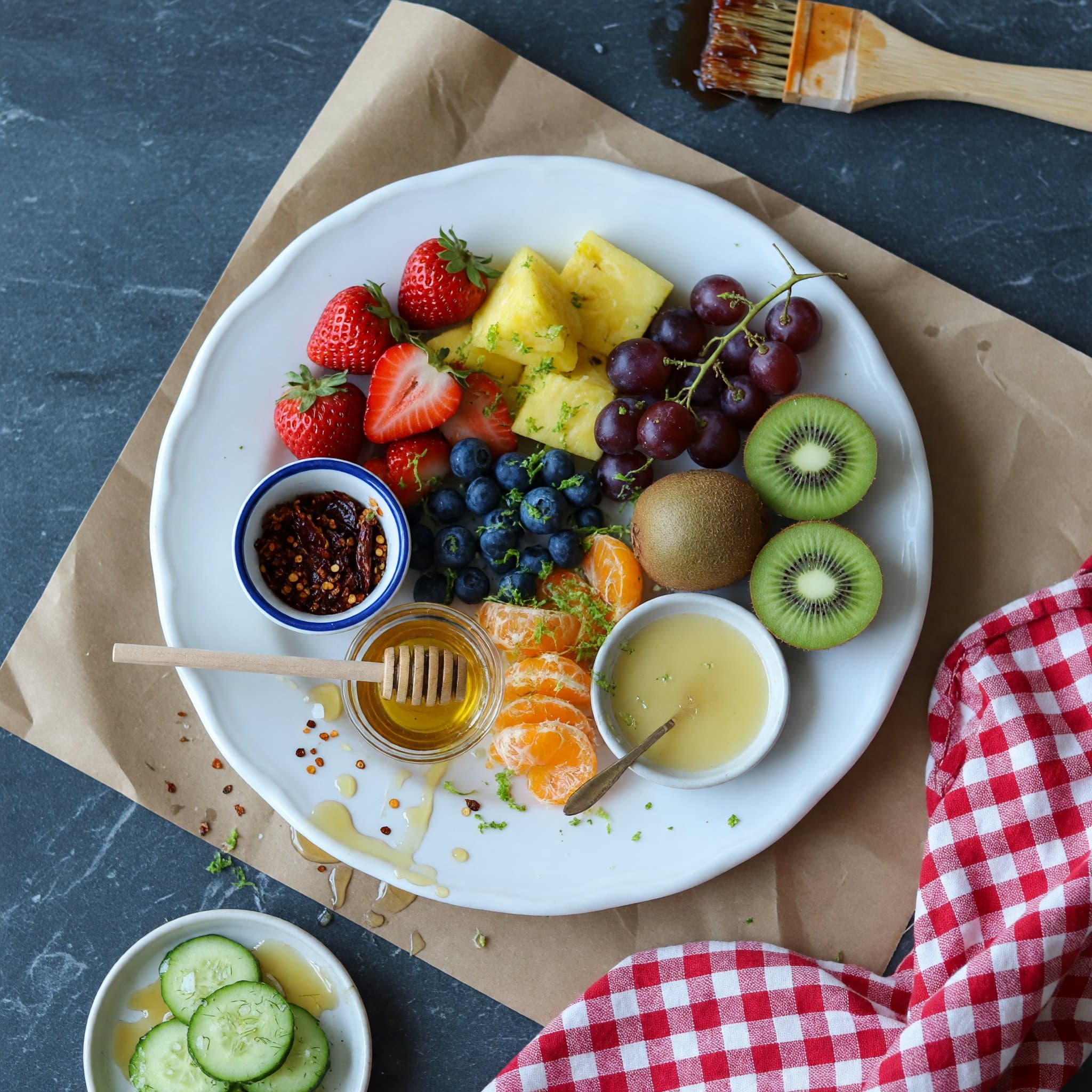 Overhead view of Strawberries, Pineapple, Blueberries, Red Grapes, Kiwis, Mandarin Oranges, Lime Zest and Honey arranged on a table
