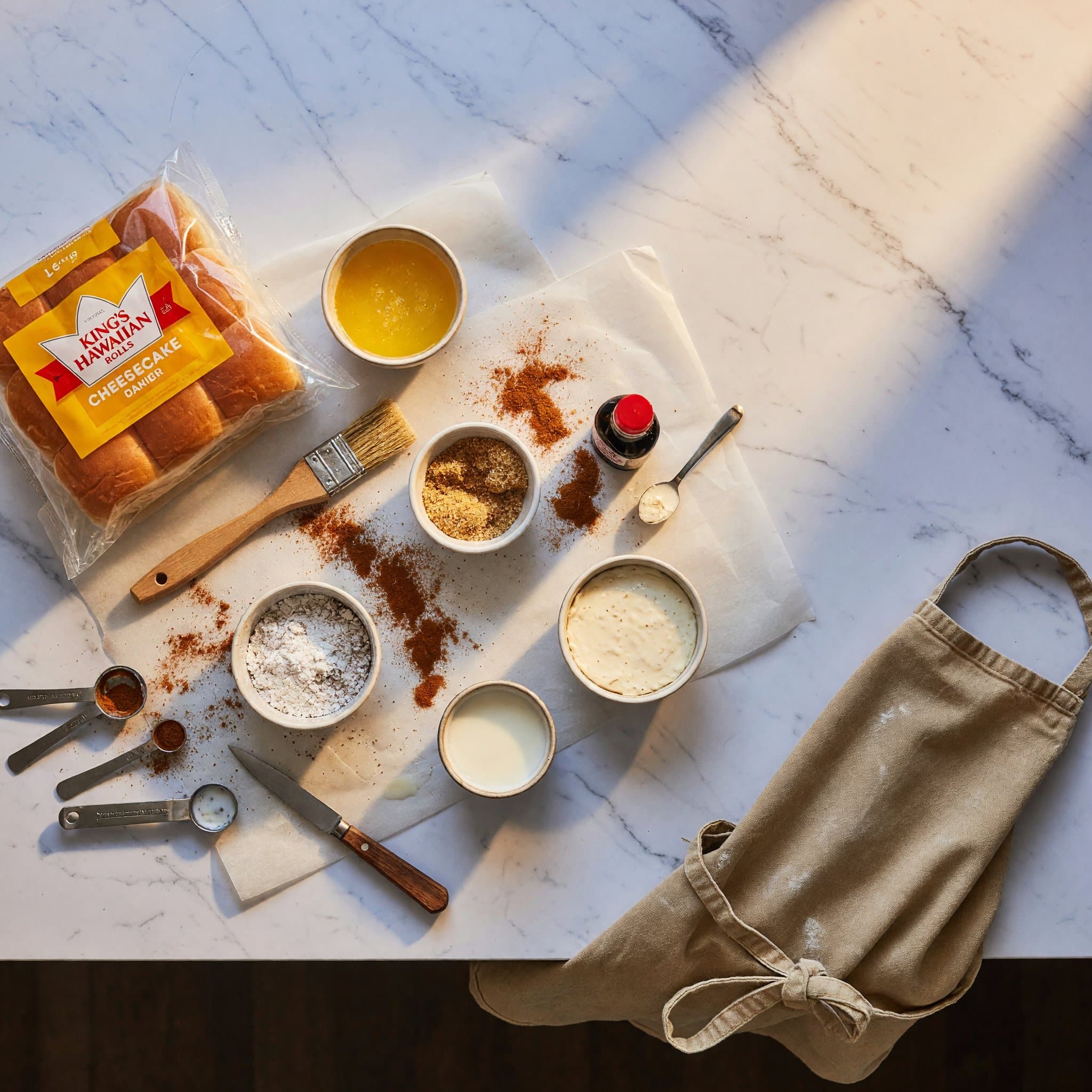 Overhead view of Hawaiian Rolls, Butter, Light Brown Sugar, Cinnamon, No Bake Cheesecake Filling, Powdered Sugar, Milk and Vanilla Extract arranged on a table